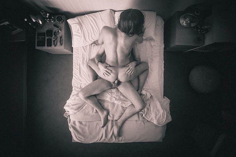 Monochrome photograph depicting a nude couple having sex in the cowgirl sex position. A slender woman astride a muscular man, their backs turned to the camera, creating an aura of privacy. Her legs are spread wide, revealing her vulva as she guides him with her hips. He grips her ass and strains against her as his erect penis penetrates her.