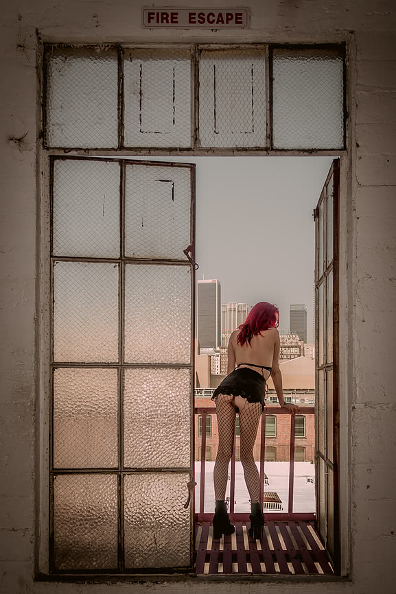 View of Lark standing naked outside in the fire escape while she looks at Downtown Los Angeles