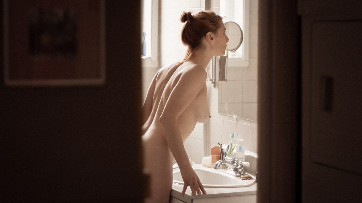 Voyeuristic view of nude blonde woman staring at herself in the mirror